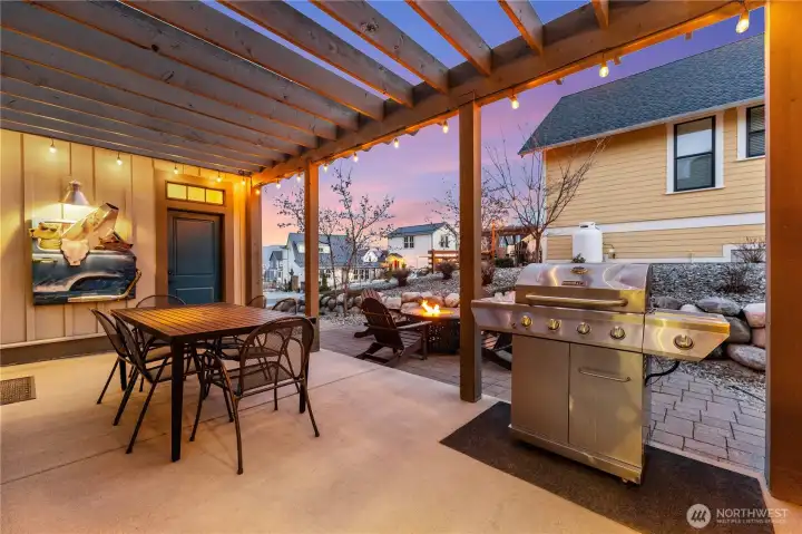 Exterior twilight view of outdoor living space with dining table, fire table with Adirondack chairs and BBQ area.