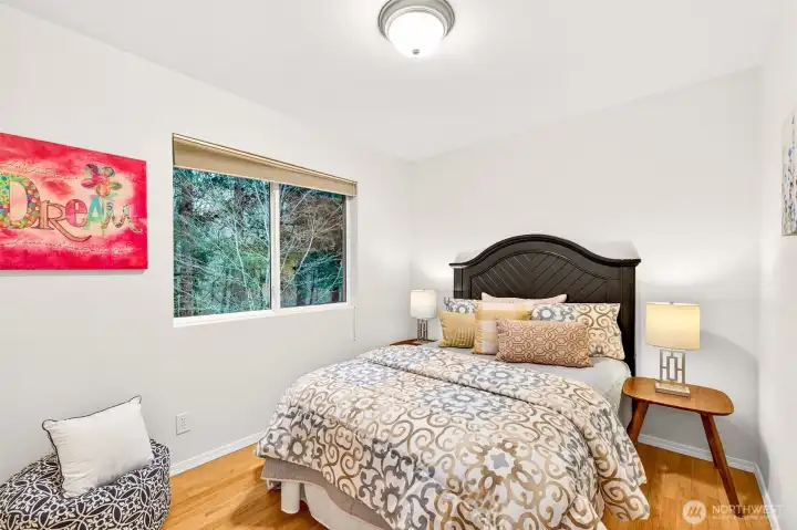 A bright and comfortable upper level bedroom with hardwood floors, fresh paint, and wooded views through a large window offers a peaceful personal space well suited as a private retreat, home office, or guest room.