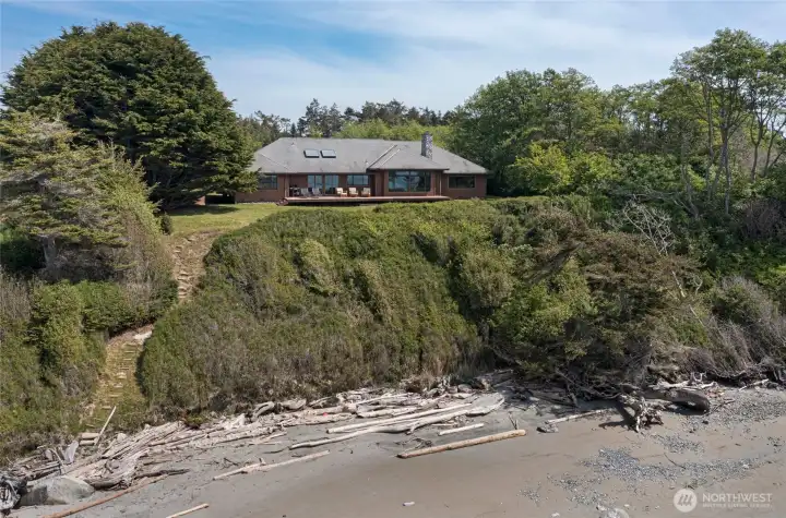The rear of the home showcases an impressive wall of large-pane windows and skylights that flood the interior with natural light, while the elevated bluff-top setting ensures unobstructed, front-row views of the glittering cove and driftwood-strewn shoreline.