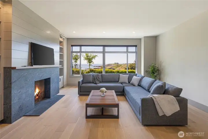 The beautifully appointed living room features a striking stone-surround fireplace, shiplap accent wall, and room for a wall-mounted TV, all framed by expansive windows showcasing breathtaking views of the San Juan Island coastline — the perfect setting for cozy evenings or entertaining guests.