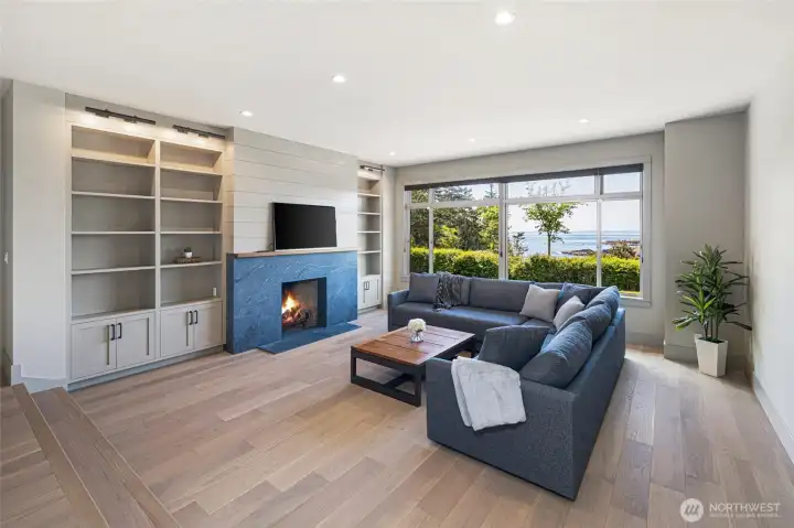 The beautifully renovated living room centers around a striking stone fireplace with shiplap surround and custom built-in shelving, while oversized windows flood the space with natural light and water views — a perfect blend of cozy warmth and coastal sophistication.