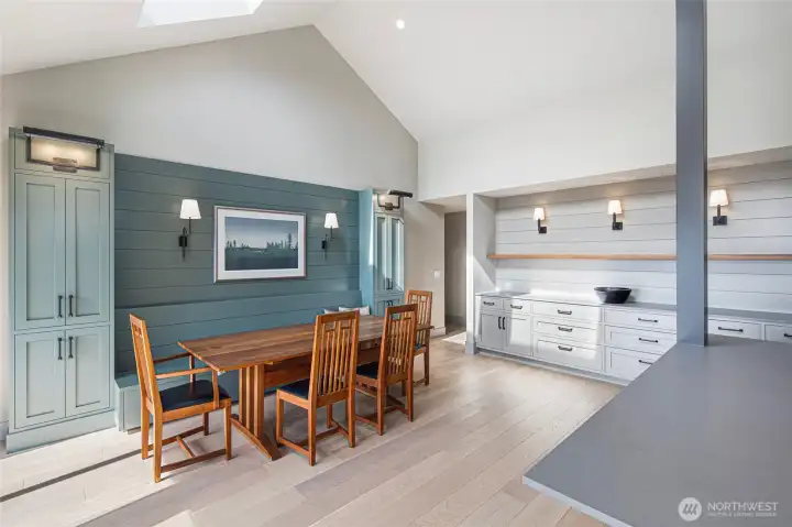A sun-drenched dining area bathed in skylight warmth is anchored by a dramatic teal shiplap feature wall and flanked by custom built-in cabinetry, creating an intimate yet airy space perfect for gathering around the table after a day on the water. The Mission-style furnishings and warm wood tones evoke a refined island sensibility.