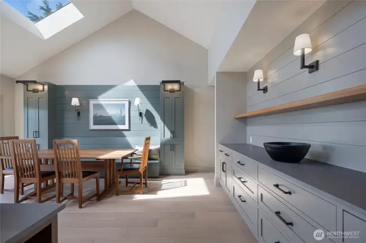 The dramatic dining area impresses with soaring vaulted ceilings, a sky-flooding skylight, custom shiplap accent walls in a signature coastal blue-green hue, built-in cabinetry, and a gorgeous craftsman dining set — the heart of island entertaining.