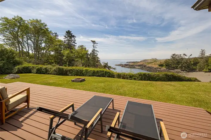 The expansive composite deck flows seamlessly into a lush manicured lawn anchored by a built-in fire pit, all framing breathtaking views of Kanaka Bay and the rocky coastline beyond. This outdoor living space is tailor-made for alfresco entertaining or peaceful sunset evenings on the island.
