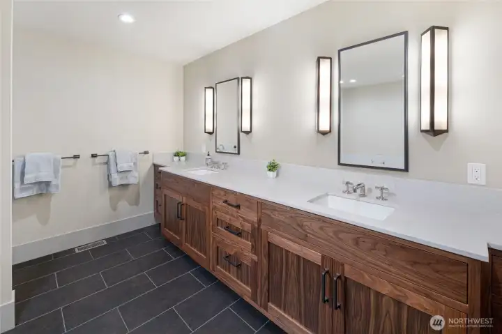 The luxurious primary bathroom features a long custom walnut double vanity topped with crisp white quartz countertops, framed mirrors with designer sconce lighting, and dark slate tile flooring — a spa-inspired haven of refined island living.