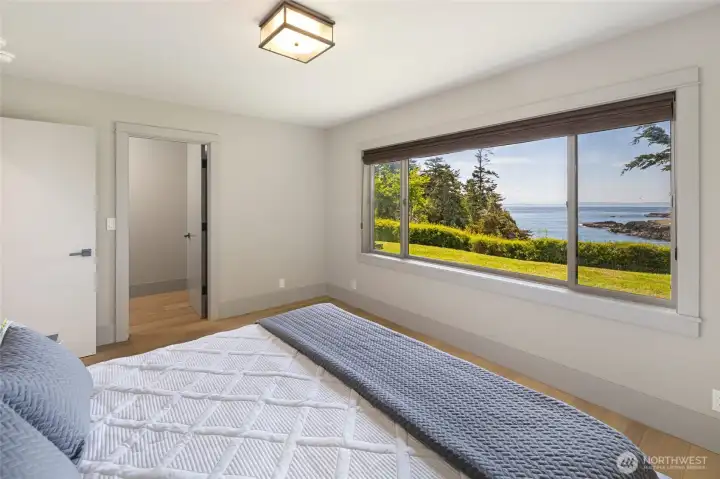 Wake up to shimmering water views from this serene guest bedroom, where a wide picture window frames the lawn, rocky shoreline, and open bay beyond in a scene that feels more like a painting than everyday life.