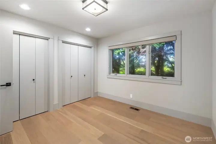 A light-filled guest bedroom offers generous dual closets, wide-plank flooring, and a triple casement window framing lush forest canopy views, providing a peaceful sanctuary for family and friends. The clean, freshly renovated palette creates a versatile space ready to be made your own.