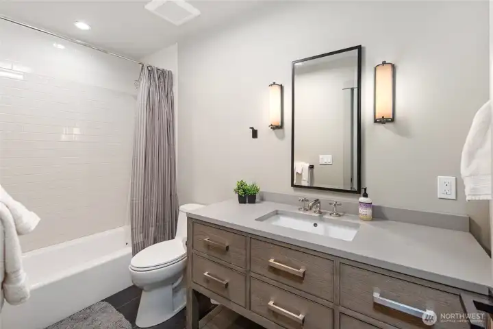 The elegantly updated full bath features a custom wood-toned vanity with quartz countertop, subway-tiled soaking tub surround, and designer black-framed mirror with warm sconce lighting for a polished, spa-like atmosphere. Clean lines and premium finishes throughout reflect the home's commitment to elevated island living.