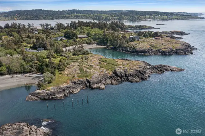 This expansive aerial captures the full drama of the property's peninsula-adjacent setting, where multiple coves, rocky headlands, and open Salish Sea waters converge in an ever-changing tableau of island scenery. The warm glow of the home's cedar exterior is visible through the trees, beckoning from its prime bluff-top perch.