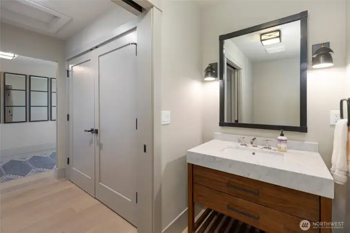 A charming powder room near the entryway showcases a warm walnut vanity topped with Carrara marble, a bold black-framed mirror, and classic sconce lighting — a thoughtful and stylish touch for guests.