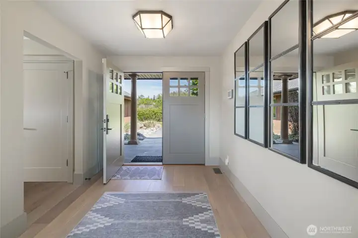 The welcoming foyer sets the tone for the home's refined coastal character, featuring light hardwood floors, elegant geometric ceiling fixtures, and a stunning craftsman-style front door opening to the island's natural beauty beyond.