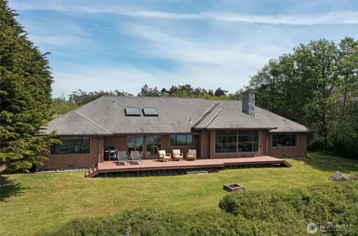 The sprawling rear deck with composite decking and comfortable seating invites effortless indoor-outdoor living, framed by towering evergreens and overlooking a lush lawn that gently gives way to the dramatic bluff and sparkling waters below.