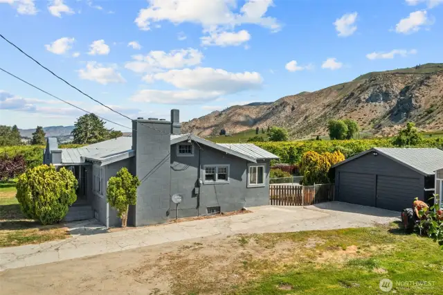 Cozy gray single-story home with detached garage, set against stunning mountain views and lush greenery.