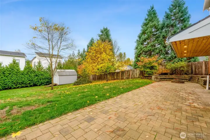 Fully fenced backyard with an extended paver patio, fire pit, and storage shed. Surrounded by mature trees for privacy and year-round greenery.
