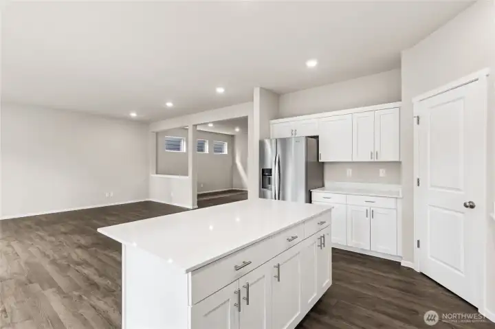 Wide-angle view of the open-concept kitchen featuring a large white quartz island, shaker cabinets, stainless steel appliances, hardwood floors, and recessed lighting with views into the great room. Photos are representative only and may display different color packages and/or upgrades.