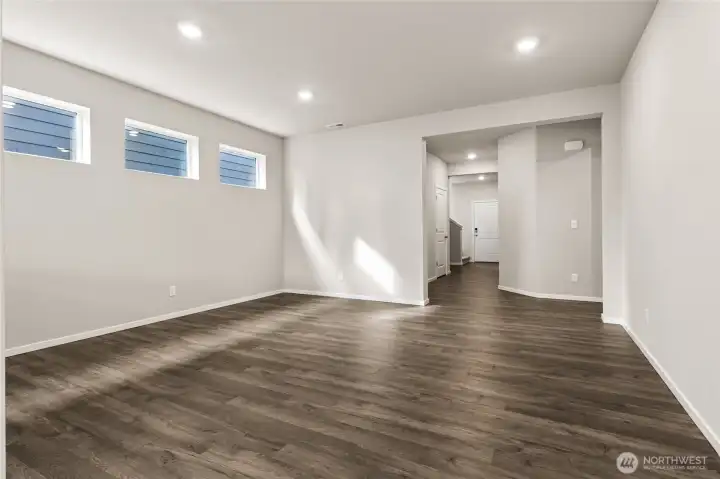 Spacious great room featuring dark hardwood floors, neutral gray walls, two windows with horizontal blinds, recessed lighting, and an open view to the hallway. Photos are representative only and may display different color packages and/or upgrades.