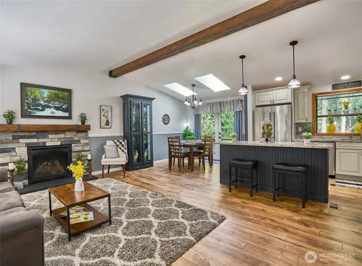 Bright, open living area with vaulted ceilings, gas fireplace with natural stone surround, matching mantel, and wood beam separating the living and dining spaces.