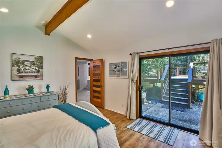 Spacious lower-level bedroom with vaulted ceilings, wood beam, laminate hardwood flooring, and a mini-split heat pump for year-round comfort. Large sliding glass door opens to the backyard and overlooks the lush yard and garden space.