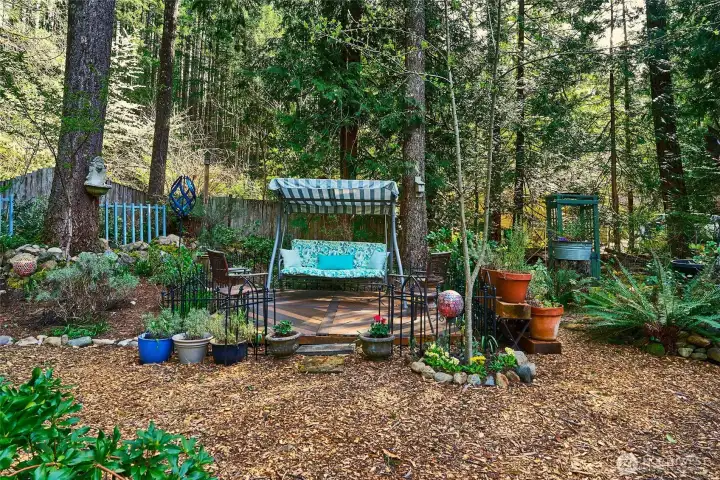 Charming wooden platform with a swinging chair nestled among lush greenery and colorful flowers—a peaceful spot to relax and enjoy the natural setting.