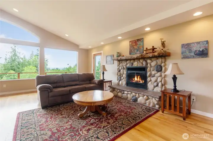 Rich hardwood floors and a striking cultured river rock fireplace with a gas insert create a warm and inviting focal point for the room. Thoughtfully designed for both comfort and functionality, the living area also features a custom-built entertainment cabinet, discreetly concealed behind a decorative wall hanging, providing organized storage for media equipment while maintaining the room's clean, attractive appearance. Large windows fill the space with natural light and showcase the beautiful views beyond.