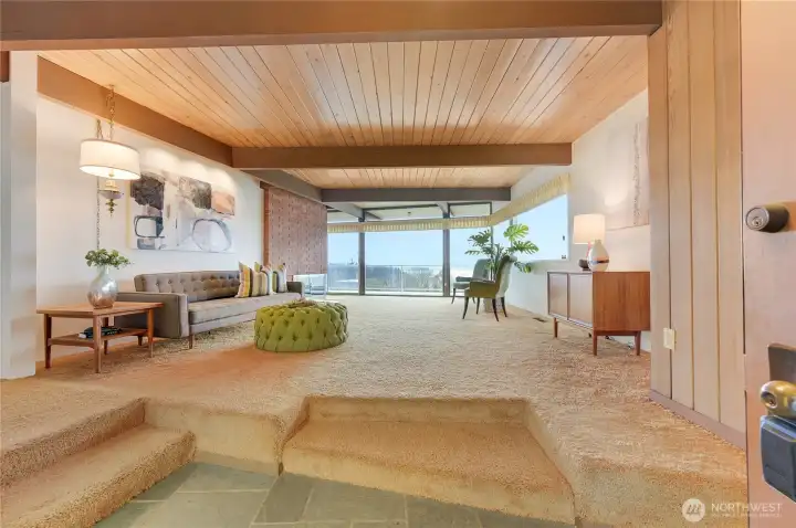 This spacious living room captures the essence of Northwest modern design, framed by sweeping views of Puget Sound and the Olympic Mountains. Warm wood ceilings with exposed beams, expansive floor-to-ceiling windows, and a seamless indoor-outdoor connection create a serene backdrop for everyday living and effortless entertaining. Natural light pours across the cozy seating area, highlighting the room’s timeless architectural character and inviting atmosphere. A rare blend of iconic design and breathtaking vista