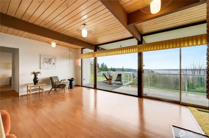 This bright and airy dining room is anchored by a warm wood-beam ceiling and expansive floor-to-ceiling glass doors that open to serene views of lush greenery and the water beyond. Sunlight fills the space throughout the day, creating an inviting atmosphere for both everyday living and effortless entertaining. The dining area flows naturally into the kitchen, enhancing functionality and connection while maintaining a beautiful sense of openness. A timeless Northwest setting designed for comfort and style.
