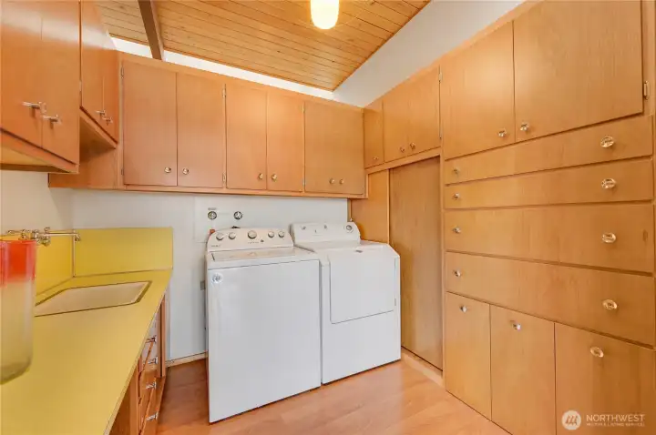 Well-appointed laundry room offers exceptional storage and everyday convenience with its extensive cabinetry, generous counter space, and built-in utility sink. Warm wood finishes and a paneled ceiling create a clean, cohesive look, while the full-size washer and dryer sit neatly within a highly functional layout. Thoughtful organization and timeless design make this a practical and inviting workspace for household tasks.