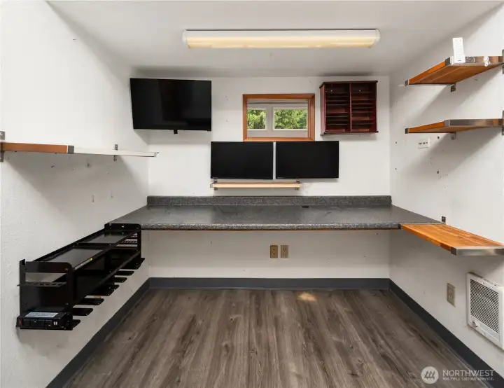 This dedicated room features an inviting work area with a  long countertop, inclusive wall-mounted monitors, storage shelves, and stylish plank style flooring.