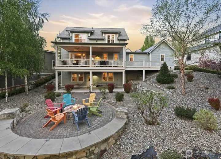 Exterior twilight view of west-facing outdoor living area with two level decks and custom stone fire pit with colorful Adirondack chairs overlooking the lake.