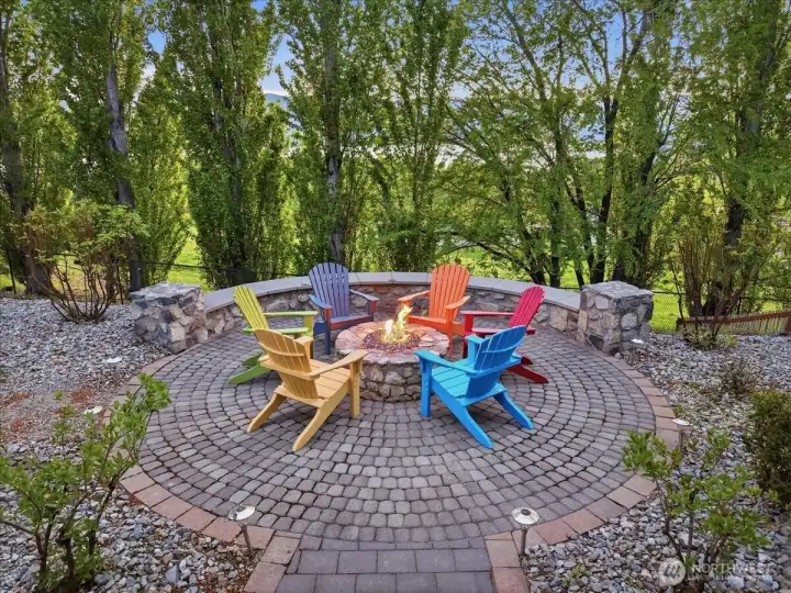 View of west-facing outdoor living area with built in stone patio and fireplace and Adirondack chairs.
