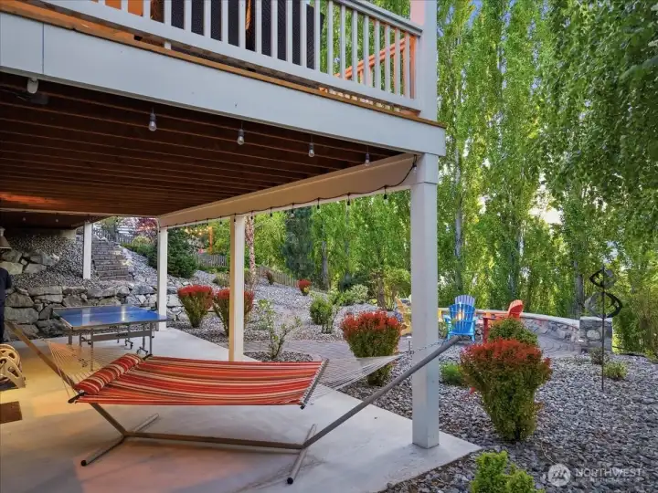 Exterior view of lower level outdoor living area with hammock, ping pong table and custom built-in stone fireplace with Adirondack chairs overlooking Lake Chelan.