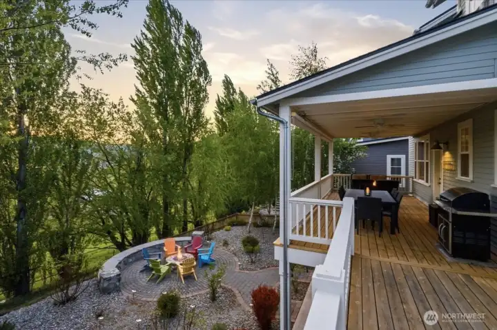 Exterior twilight view of outdoor living area with main level lake view deck with dining area, BBQ and fire table/lounge area and lower level with custom built-in stone fireplace and Adirondack chairs.