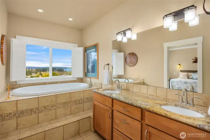 Primary ensuite bath with double vanity, granite countertops, and soaking tub beneath a window capturing beautiful water views.