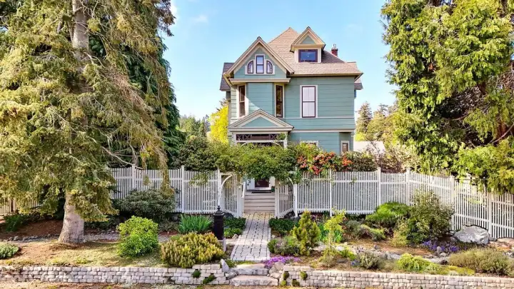 An 1889 Victorian with distive gables, detailed trim, and a covered entry reached through a white picket gate and established gardens.