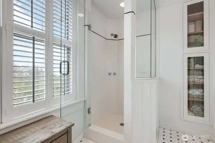 An updated shower alongside glass-front cabinetry and shuttered windows, with natural light throughout.