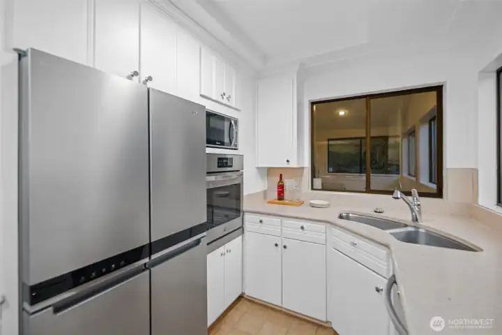Beautifully integrated stainless steel appliances anchor this part of the kitchen, including the double door refrigerator, elevated oven, and built in microwave. A wide window above the double sink brings in soft natural light and another looks into the sunroom, adding a warm sense of connection. Refined finishes, clean lines, an efficient use of space with generous cabinetry, all create a bright workspace that feels both elegant and thoughtfully designed.