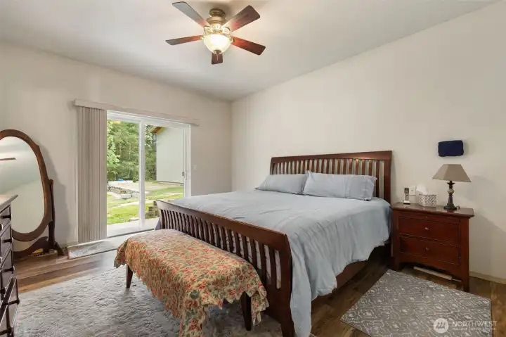 The primary bedroom offers a spacious retreat with warm wood grain laminate floors, room for substantial furniture, and direct access to the back patio with views toward the trees, yard, and outdoor spaces.