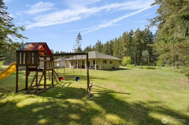 A backyard built for real life, with room to play, explore, gather, and grow, all wrapped in green lawn, trees, and a glimpse of the pond beyond.