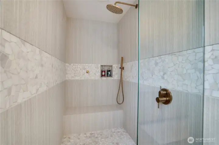 The walk-in shower features a custom marble mosaic accent band, built-in bench, rain head, hand wand, and frameless glass enclosure — finished in brushed gold. The walk-in shower features a custom marble mosaic accent band, built-in bench, rain head, hand wand, and frameless glass enclosure — finished in brushed gold.