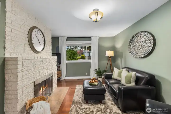 Additional living room with painted brick fireplace, hardwood floors, and large window for natural light. This comfortable secondary living space offers a cozy setting for relaxing, reading, or flexible everyday use.