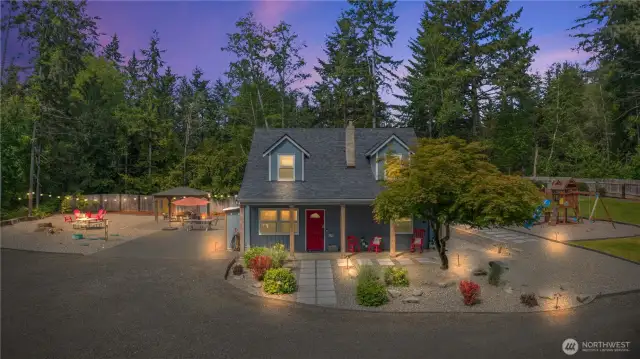 Experience the magic of this storybook home at twilight! This beautifully lit exterior highlights a vibrant red front door, charming covered porch, and inviting seating area under the soft glow of outdoor lighting. To the left, a cozy fire pit and gazebo create the ultimate backyard entertaining zone, while the right side reveals a fully fenced play area—perfect for children or pets. Surrounded by majestic evergreens, this serene oasis offers privacy, peace, and Pacific Northwest charm.