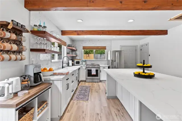 This full view of the kitchen showcases the oversized quartz island with breakfast bar seating, perfect for casual dining or entertaining guests. Stainless steel appliances, sleek black hardware, and a smart layout provide both style and function. The open shelving and custom mug rack bring warmth and character to this show-stopping culinary space.