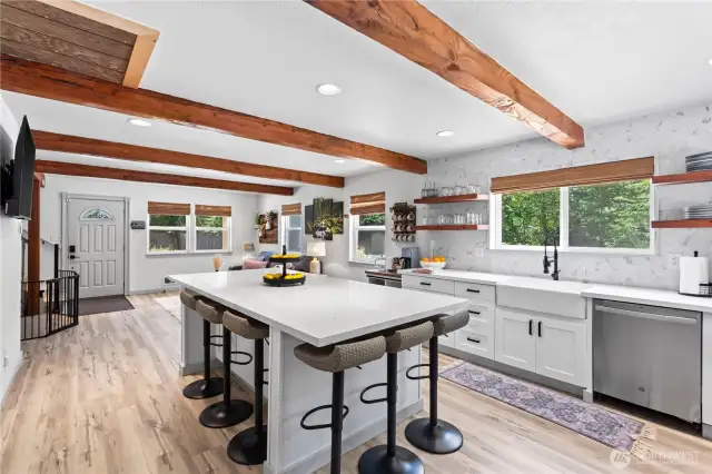Designed with gathering in mind, this angle highlights the seamless flow from kitchen to living area. The expansive island with bar stools is the heart of the home, complemented by textured wood beams, wide-plank flooring, and a backdrop of open shelving and abundant windows. This layout invites you to cook, connect, and relax all in one place.