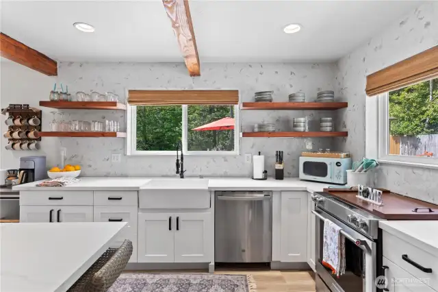 A closer look at the kitchen’s heart reveals a deep farmhouse sink paired with sleek open shelving, matte black fixtures, and a marble-style backsplash. The mix of textures and thoughtful design adds both elegance and utility. Stainless steel appliances, including a dishwasher and gas range, complete the polished look.