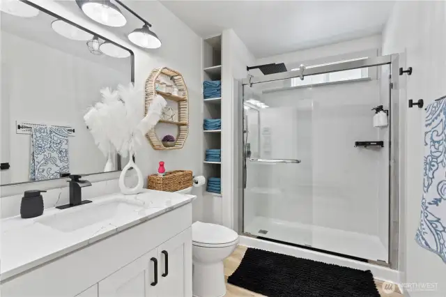 This beautifully remodeled bathroom offers a spa-like experience with a sleek walk-in shower, quartz countertop, matte black fixtures, and built-in shelving for added storage. Woven textures, soft lighting, and modern finishes blend to create a stylish yet functional space that feels fresh and luxurious.
