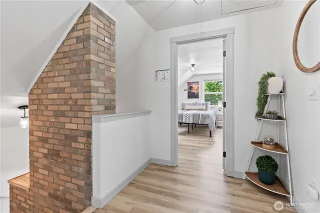 This inviting upstairs hallway features a charming brick accent chimney and leads into a bright, spacious bedroom. The open walkway is adorned with thoughtful decorative shelving and warm-toned flooring, creating a natural transition between the home’s upper-level living spaces.