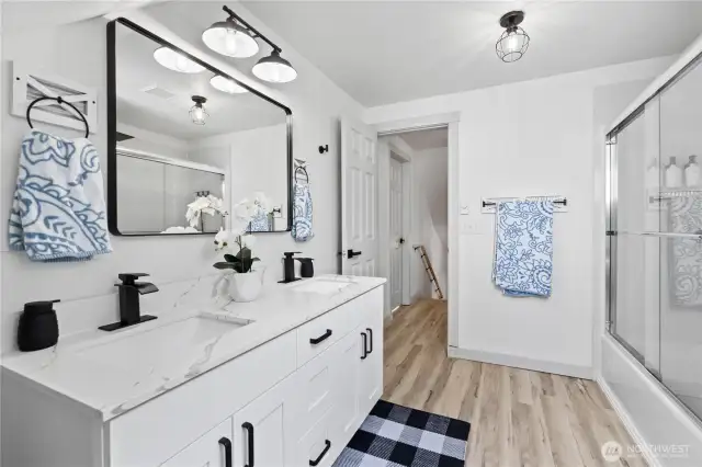 This beautifully updated bathroom features a dual-sink vanity with elegant quartz countertops, matte black fixtures, and a full tub/shower combo with sliding glass doors. The generous layout and thoughtful storage provide both form and function—perfect for shared use or guests.