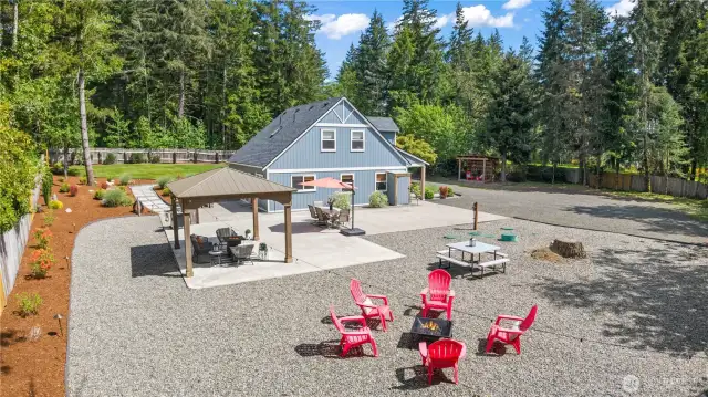 Aerial drone view of a stunning property that blends modern charm with ultimate functionality. The expansive backyard features a gravel fire pit area with red Adirondack chairs, a large concrete patio with multiple seating zones, and a beautifully maintained blue farmhouse-style home. The spacious layout includes manicured landscaping, room for RV parking, and ample space for gatherings or future projects.