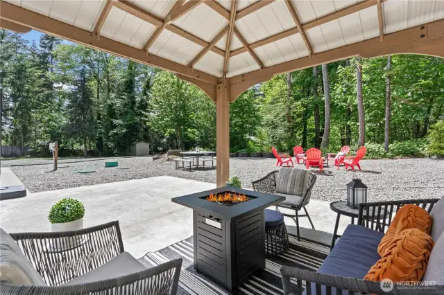 Chic and cozy outdoor living at its finest! This covered gazebo features a stylish gas fire table surrounded by modern cushioned seating, creating the perfect hangout for chilly evenings or casual afternoons. The open view reveals a fire pit area and picnic table, offering multiple zones for relaxing and entertaining. Surrounded by evergreens and designed for comfort, this setup transforms any backyard into a peaceful retreat.