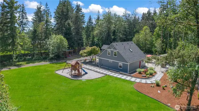 The perfect blend of nature and recreation! This wide-angle view highlights a meticulously maintained lawn, custom stone pathway, and a well-positioned play structure, all surrounded by lush evergreens. The side of the home features additional landscaped areas, offering both function and beauty. Whether you envision kids playing freely or hosting weekend gatherings, this space delivers flexibility, fun, and a peaceful backdrop.